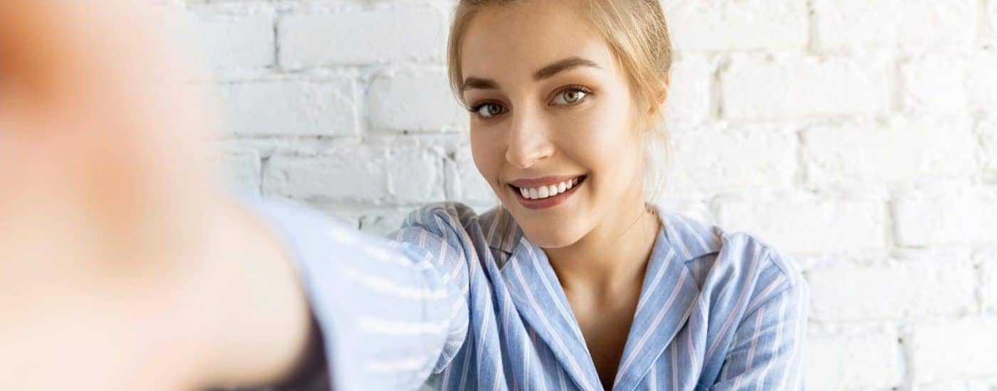 Portrait of attractive young woman in striped pyjamas making video call. Lovely model posing on white brick wall at home. Technology and good morning concept.