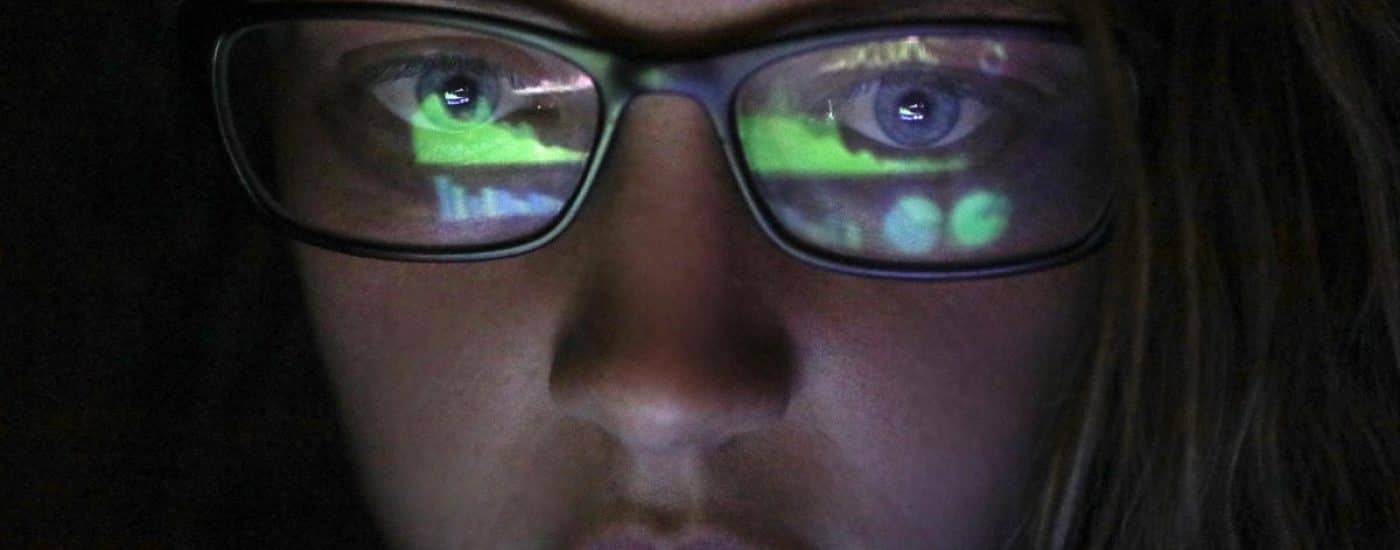 an Adult female looking at financial graphs on a laptop in a dark room with financial graphs reflected in his glasses.